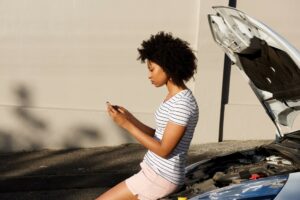 A woman sits on the bonnet of her white car after it has suffered a mechanical breakdown. She phones her insurance provider to tow her car to the nearest approved mechanic according to her vehicle motor warranty.
